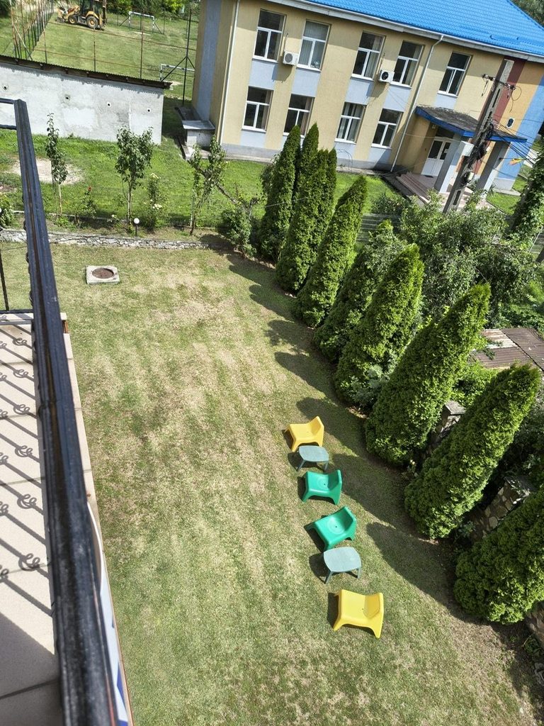 Priveliștea peste gazonul curții, șirul de tuia și scaunele colorate de pe iarbă