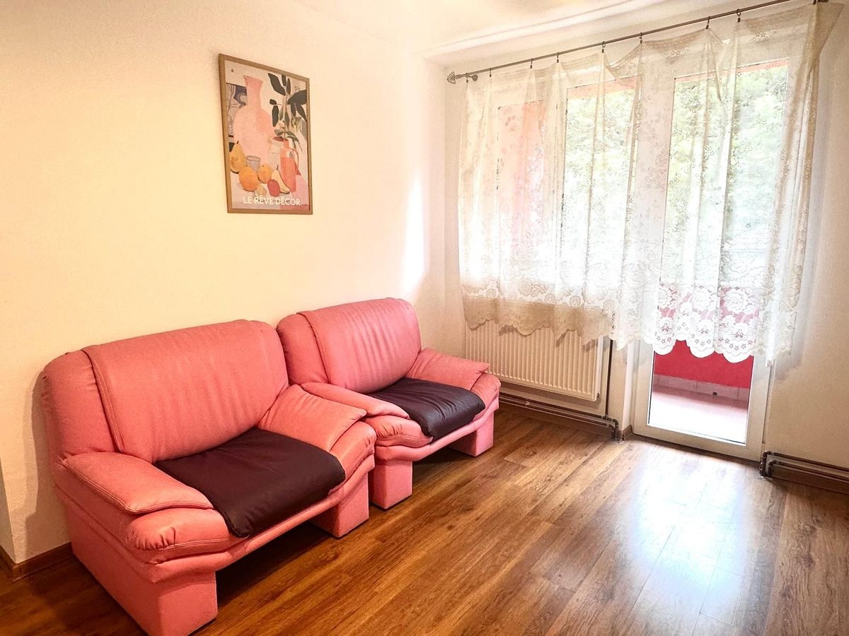 Camera de zi a unui apartament, cu canapea și fotoliu roz, parchet și ușă spre afară