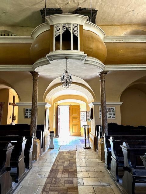 Nava bisericii catolice privită dinspre altar, cu balconul orgii deasupra intrării și ușa deschisă spre lumina de afară