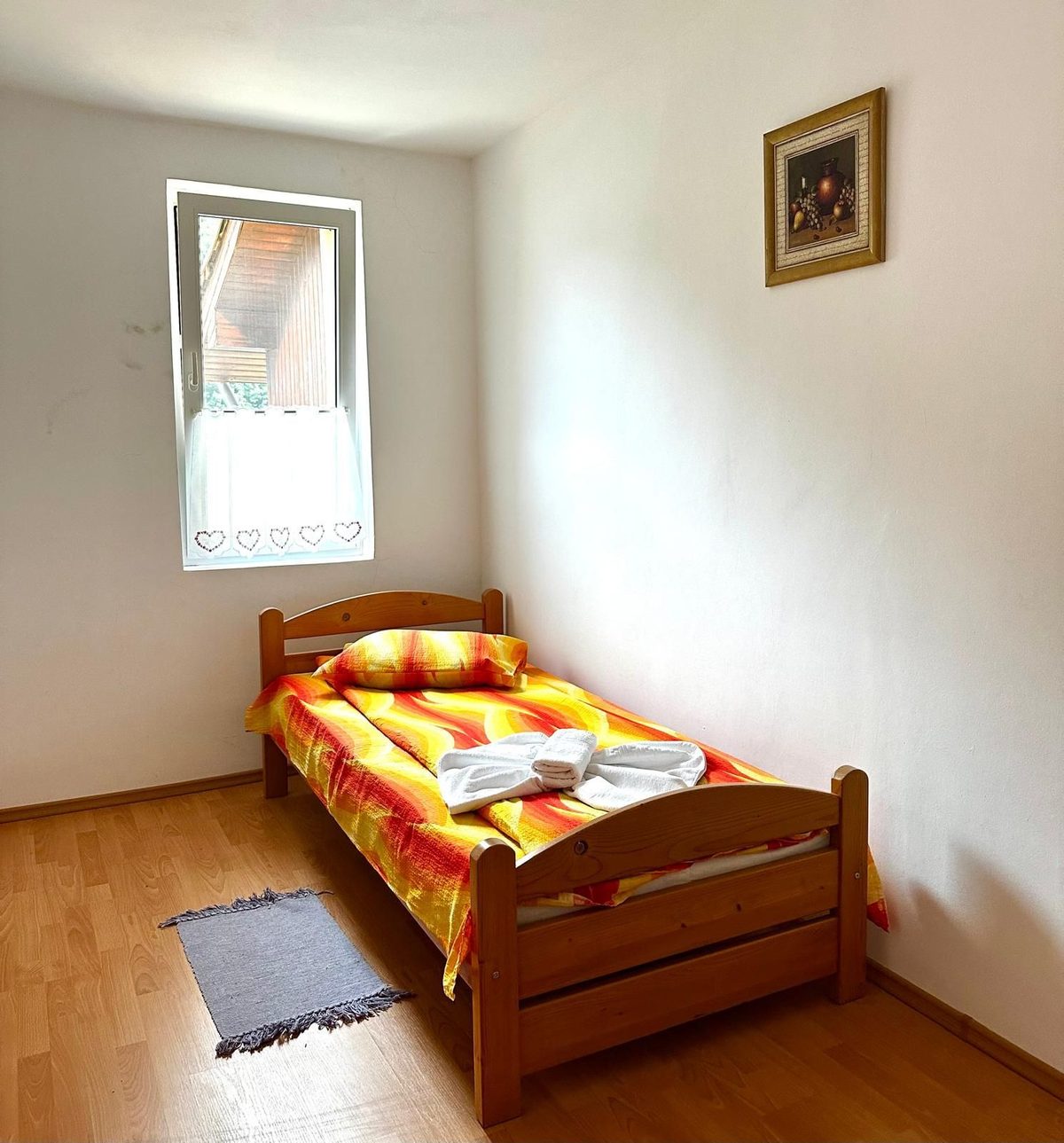 Camera single dintr-un apartament, cu pat de brad, cuvertură galbenă și perdea brodată cu inimi