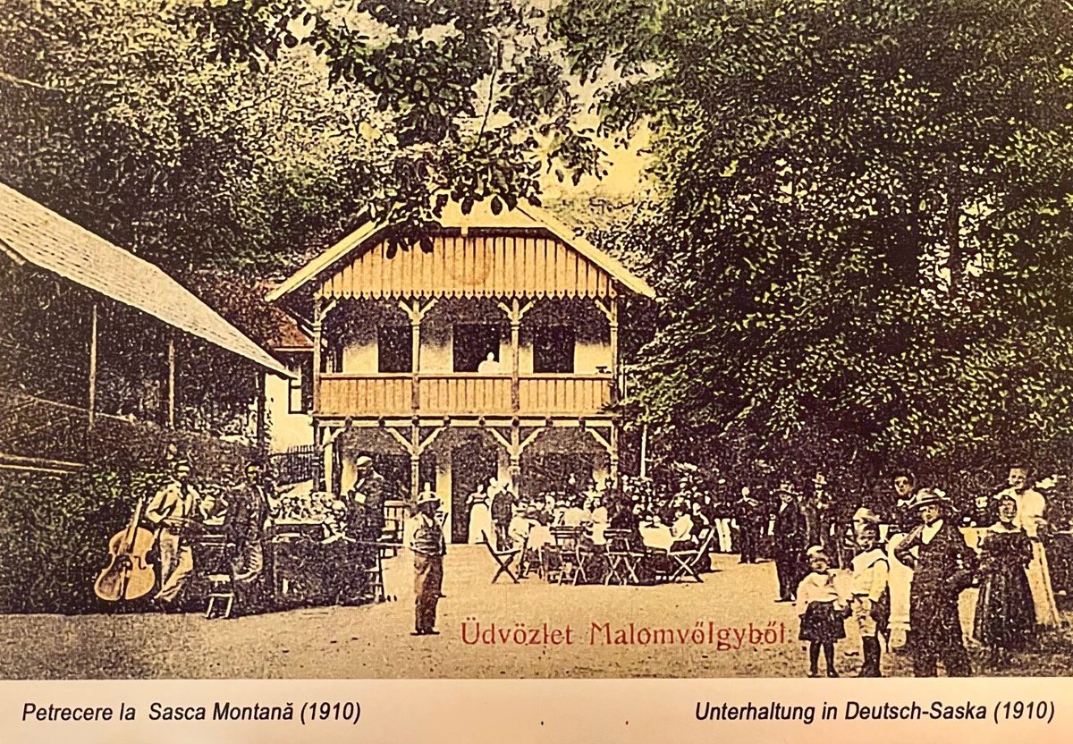 Carte poștală din 1910: lume la mese sub cerdacul de lemn al clădirii din Valea Morii, cu doi lăutari cu contrabas în stânga și copii îmbrăcați de duminică în față