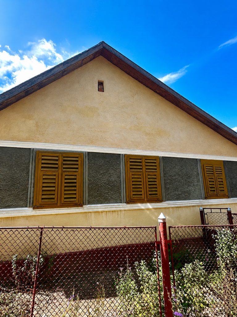 Casă bănățeană cu fața la drum, cu patru obloane de lemn galben și soclu vopsit gri, sub un cer de vară fără nori