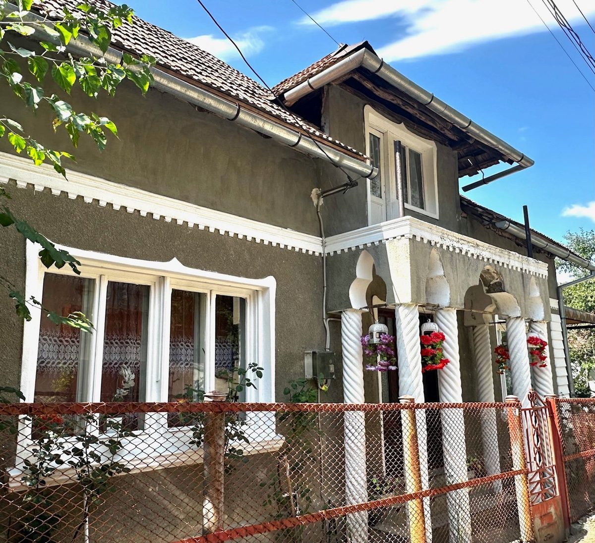 Casă cu pridvor închis și coloane albe, cu ferestre cu perdele dantelate și un gard roșu de plasă în față