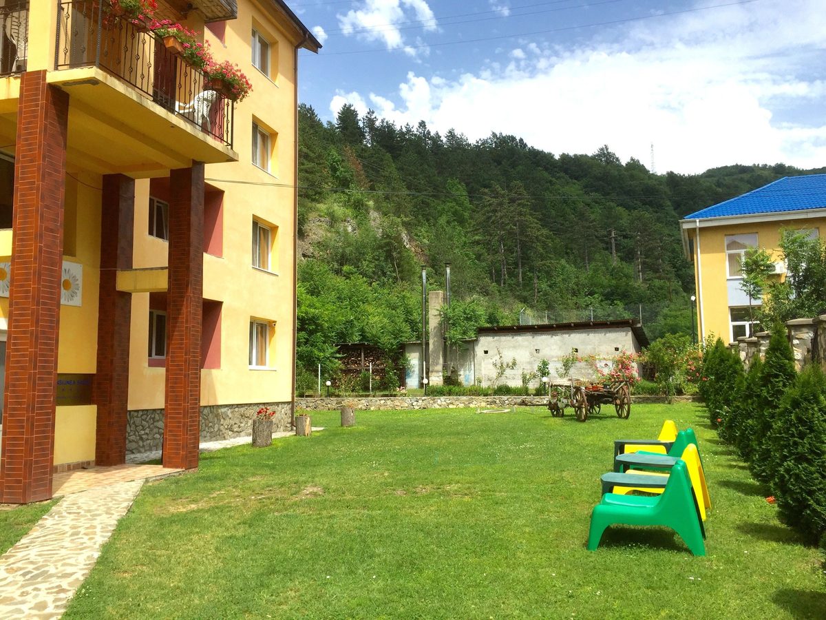Curtea pensiunii primăvara, cu iarbă tunsă și dealul împădurit deasupra