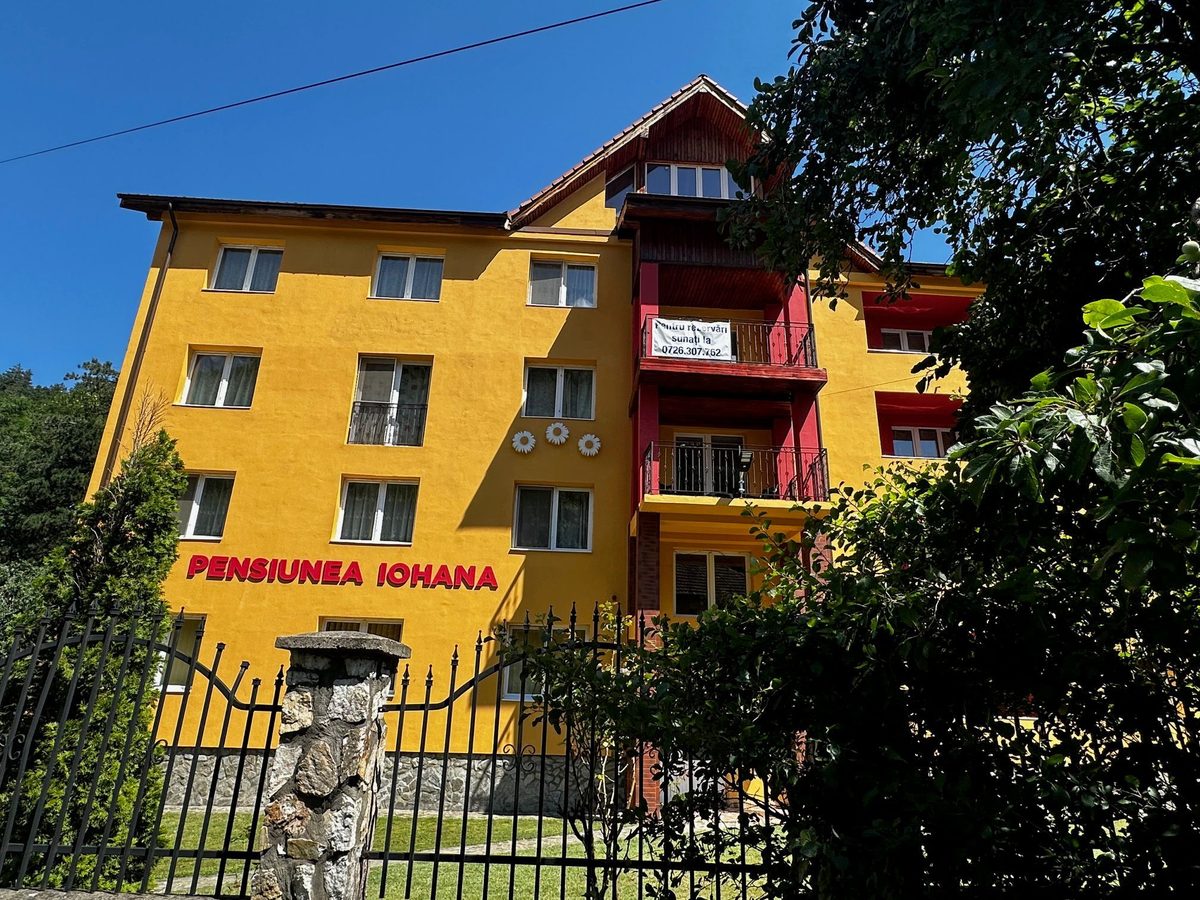 Clădirea pensiunii văzută de la poarta de fier forjat, peste gardul de piatră de râu