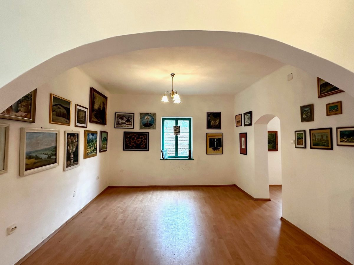 Galeria văzută pe sub arc, cu tablouri pe toți pereții și lumina intrând prin fereastra turcoaz