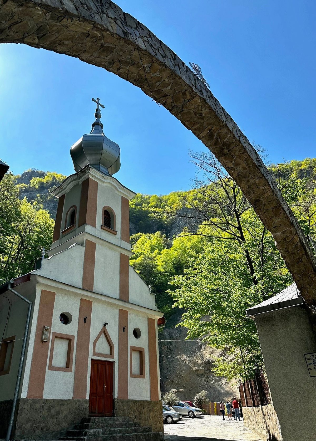 Biserica Mănăstirii Călugăra văzută pe sub arcul de zidărie de la intrarea în incintă, cu versantul împădurit deasupra