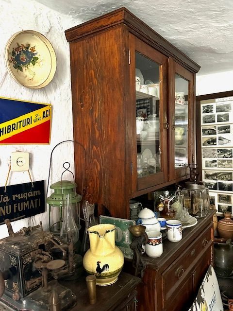 Vitrină de lemn cu vase de porțelan, între o tablă emailată „Chibrituri se vând aci” și una cu „Vă rugăm nu fumați”