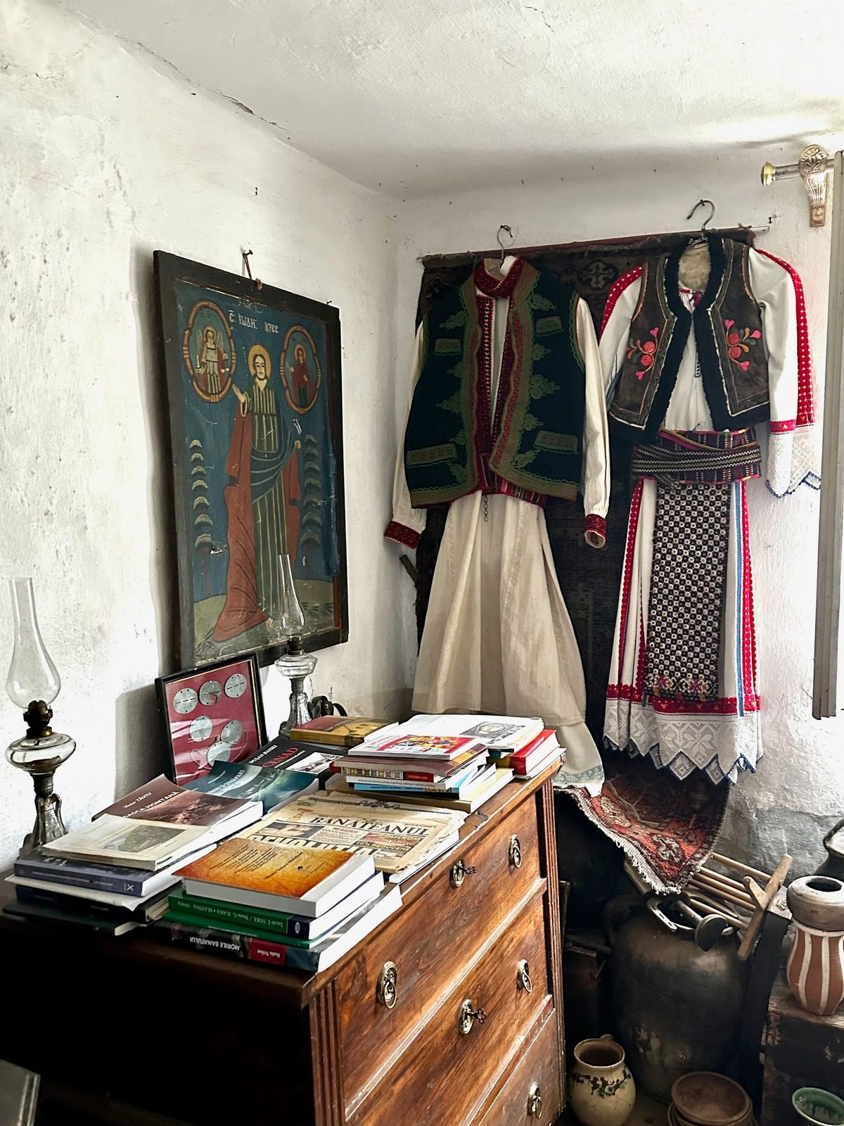 Colț de cameră din muzeu, cu o icoană pictată pe lemn, două costume populare pe umeraș și o comodă încărcată de cărți despre Banat