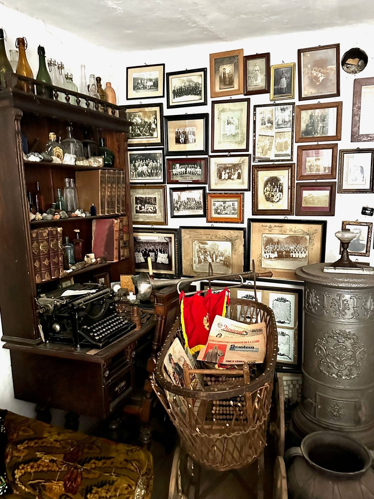 Perete acoperit din tavan până jos cu fotografii de familie înrămate din Sasca de la 1900, lângă o mașină de scris veche și o sobă de fontă