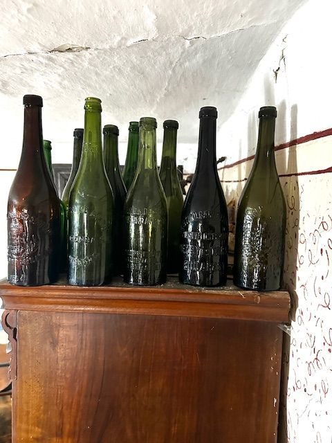 Sticle de bere din secolul trecut, cu numele fabricii ștanțat în sticla verde și brună, așezate pe o comodă